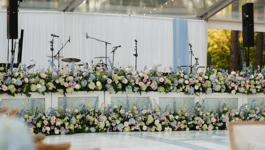 Lost Wax stage set with drums and microphones behind a flower-lined dance floor in the reception tent at Grace + Heath's wedding in Texarkana, TX in 2025